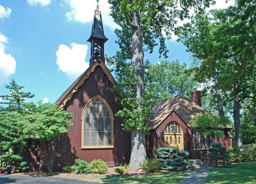 A charming wooden church with a tall steeple, surrounded by lush greenery and blue skies.