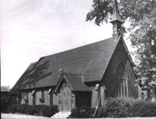 A historic wooden church with a steep roof, pointed steeple, and decorative entrance, surrounded by trees and shrubs.