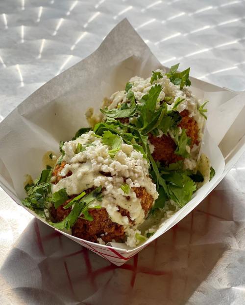 Two crispy, golden-brown fritters topped with fresh herbs and drizzled with creamy sauce, served in a paper tray.