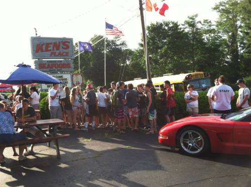 A crowd of people waiting outside Ken's Place seafood restaurant, with a red sports car parked nearby.