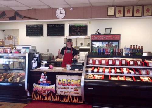 A smiling employee stands behind a counter in a BBQ restaurant, surrounded by food displays and a welcome sign.
