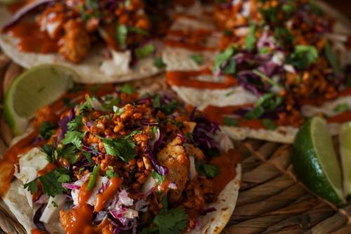 Three tacos topped with shredded cabbage, cilantro, and spicy sauce, served with lime wedges on a woven platter.
