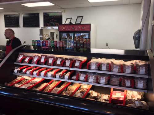 A meat display case filled with various packaged meats, with a butcher in the background and drinks on a shelf.