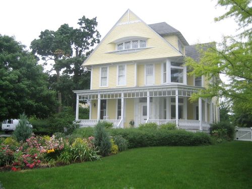 A large yellow Victorian house with a wraparound porch, surrounded by lush greenery and colorful flower beds.