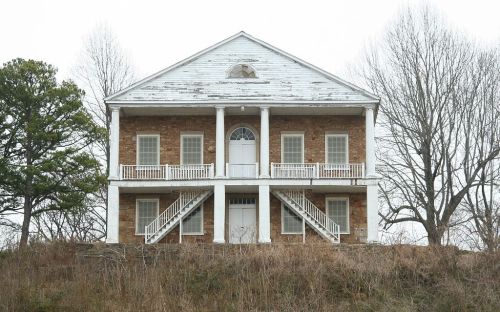 A two-story brick house with white columns and stairs, surrounded by bare trees and overgrown grass.