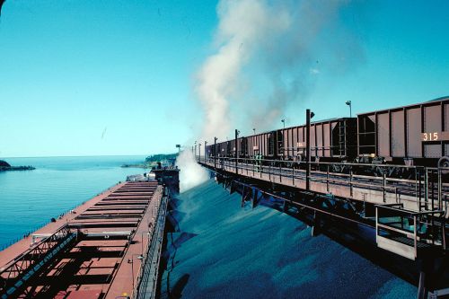 A cargo ship loading coal from train cars by the water, with smoke rising against a clear blue sky.