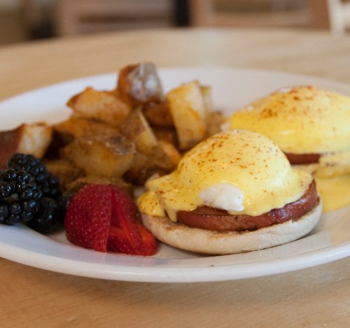 A plate of eggs Benedict with ham, hollandaise sauce, and a side of roasted potatoes, strawberries, and blackberries.