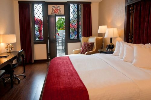 Cozy hotel room with a king bed, armchair, desk, and large windows featuring red curtains and a view of the outdoors.