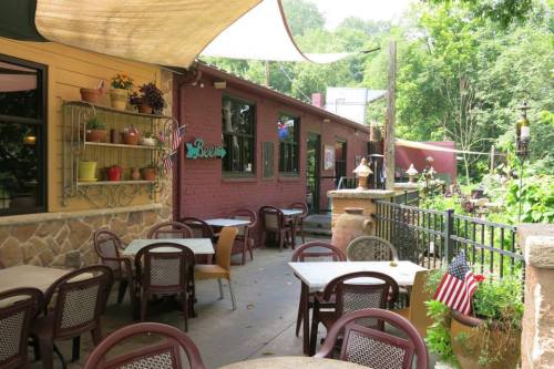 Outdoor dining area with tables, chairs, and greenery, featuring a rustic building and decorative elements.