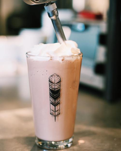 A tall glass of chocolate milkshake topped with whipped cream and a decorative feather design.