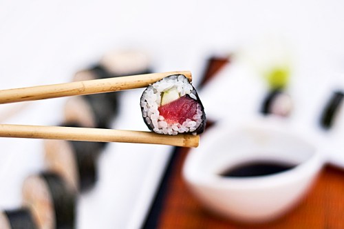 A piece of sushi held with chopsticks, featuring tuna and rice, with soy sauce in the background.