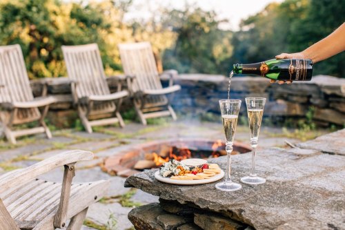 A hand pours champagne into two glasses beside a plate of cheese and fruit, near a fire pit and wooden chairs outdoors.