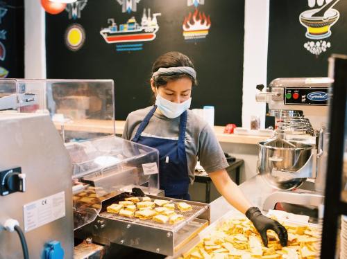 A worker in a mask prepares food in a kitchen, surrounded by cooking equipment and colorful wall art.