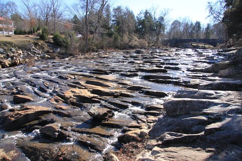 A rocky riverbed with flowing water, surrounded by trees and a bridge in the background under a clear blue sky.