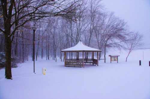 A snow-covered gazebo surrounded by bare trees in a winter landscape, with soft snowfall and a serene atmosphere.
