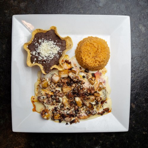 A plate featuring grilled chicken with cheese sauce, rice, and a tortilla bowl filled with black beans.