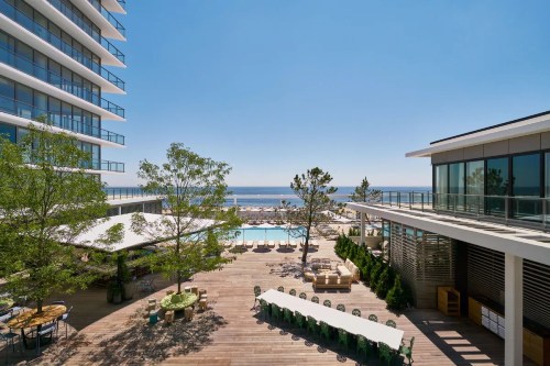 A sunny outdoor area with a pool, lounge chairs, and modern buildings overlooking the ocean.