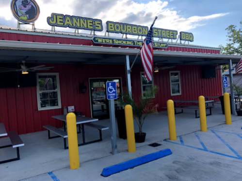 Exterior of Jeanne's Bourbon Street BBQ with red walls, picnic tables, and an American flag. Accessible parking visible.