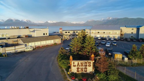 Aerial view of a coastal area with a "Lands End" sign, buildings, and mountains in the background under a clear sky.