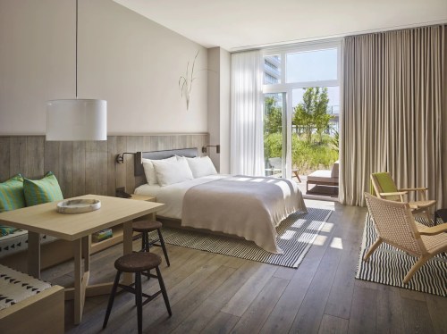 Bright, modern hotel room with a large window, wooden floors, a bed, dining table, and cozy seating area.
