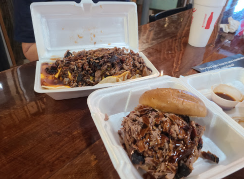 Two takeout containers with barbecue: one with pulled pork on a bun and the other with chopped pork over fries.