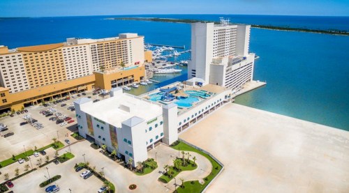 Aerial view of a coastal hotel and marina, with clear blue water and a sunny sky in the background.