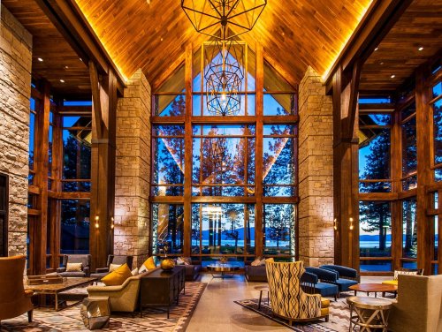 A spacious lodge interior with large windows, wooden beams, and cozy seating, showcasing a sunset view over a lake.