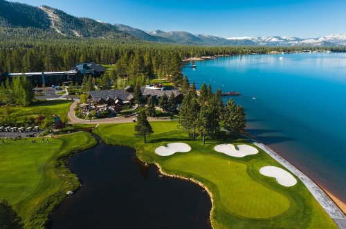 Aerial view of a scenic golf course by a lake, surrounded by trees and mountains under a clear blue sky.