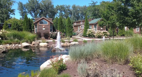 A serene landscape featuring a pond with a fountain, surrounded by greenery and rustic cabins under a clear blue sky.