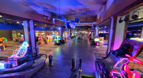 A brightly lit arcade with colorful games and neon lights, featuring an airplane model hanging from the ceiling.