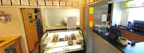 A cozy café interior featuring a display case of desserts and a counter with menus and colorful signs.