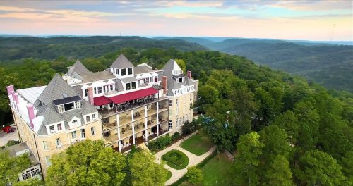 Aerial view of a grand hotel surrounded by lush green hills and a colorful sunset sky.