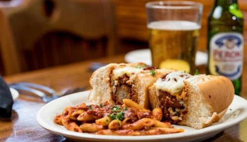 A plate with a meatball sub topped with cheese, served alongside penne pasta in marinara sauce and drinks in the background.