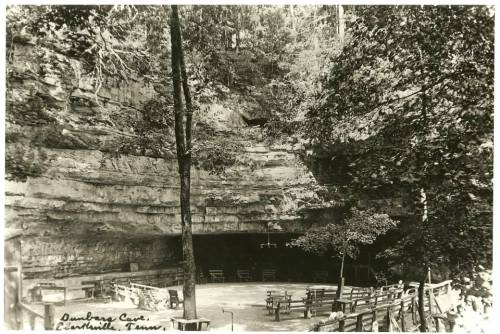 Black and white photo of a cave entrance surrounded by trees, with benches arranged in front.