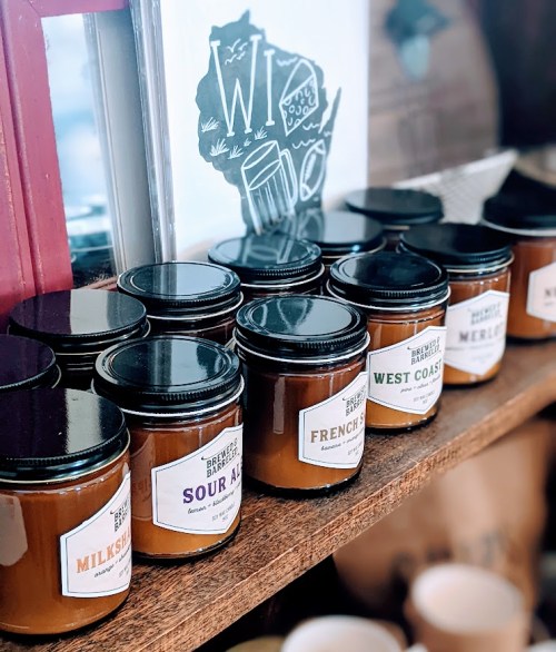 A display of various candle jars with labels, arranged on a wooden shelf in a cozy shop setting.