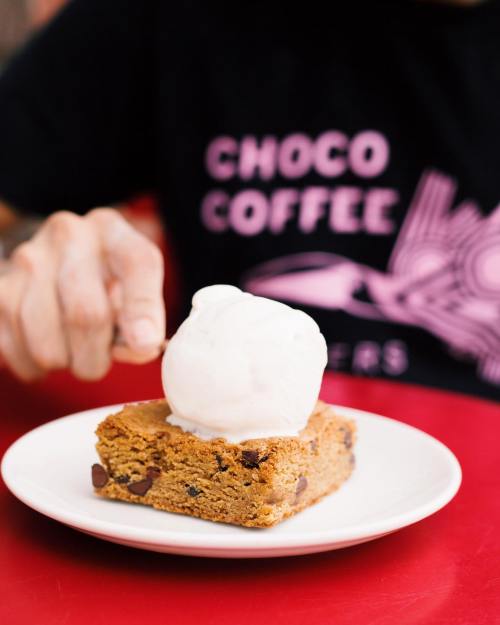 A hand reaches for a scoop of ice cream on a warm chocolate chip cookie bar on a white plate.