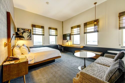 A modern hotel room featuring a bed, seating area, desk, and large windows with natural light.