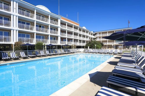 A sunny hotel pool area with striped lounge chairs and umbrellas, surrounded by a multi-story building.