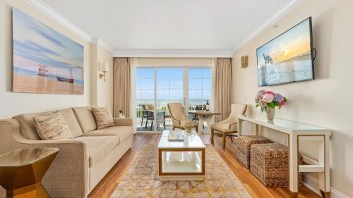 Bright and airy living room with a beige sofa, glass coffee table, and ocean view through large windows. Decorated with art.