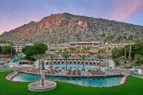 A luxurious resort with a pool, palm trees, and a mountain backdrop under a colorful sky at sunset.