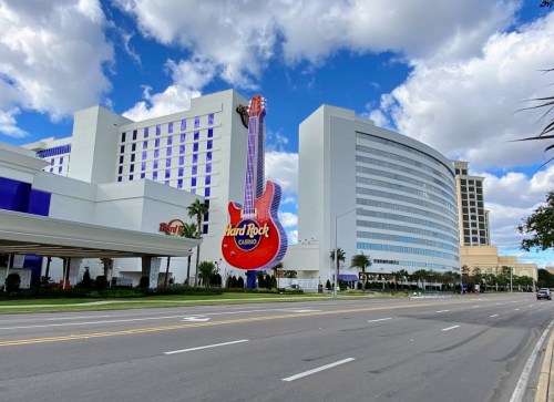 A large guitar sign for Hard Rock Casino stands in front of a modern hotel against a blue sky with clouds.