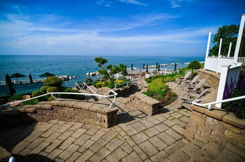 Scenic view of a lakeside patio with stone pathways, lounge chairs, and lush greenery under a blue sky.