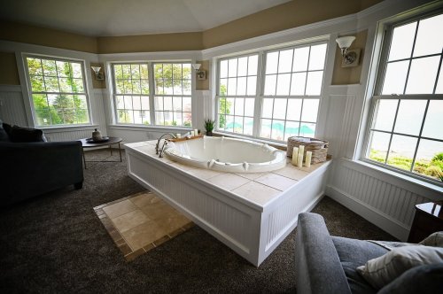 A cozy bathroom with a large tub, surrounded by windows and soft lighting, featuring a plush carpet and a comfortable chair.