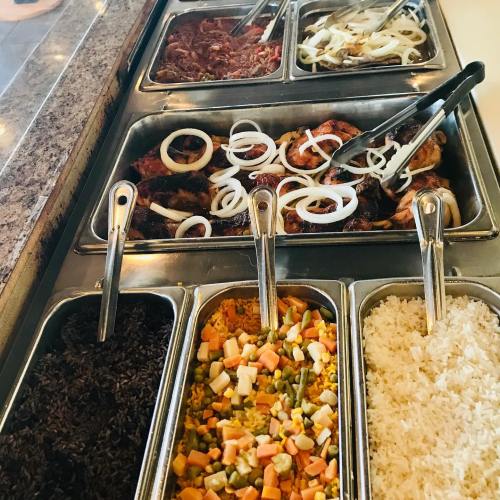 A buffet display with various meats, grilled onions, and sides of rice, mixed vegetables, and black rice in metal trays.
