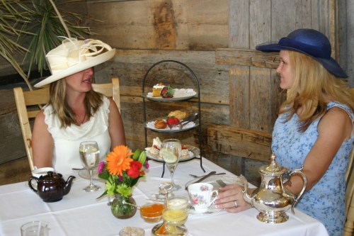 Two women in elegant hats enjoy a tea party, seated at a table with a variety of treats and drinks in a rustic setting.