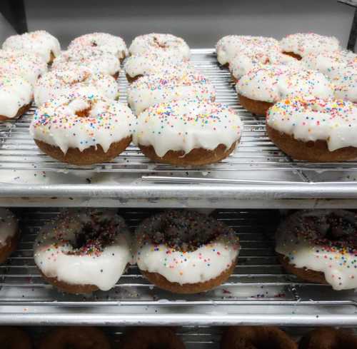 A tray of freshly baked donuts topped with white icing and colorful sprinkles, arranged on wire racks.