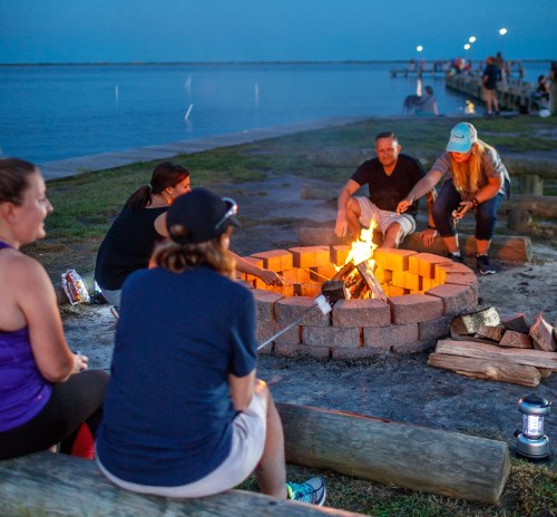 A group of people gathered around a fire pit at dusk, roasting marshmallows by a lakeside.