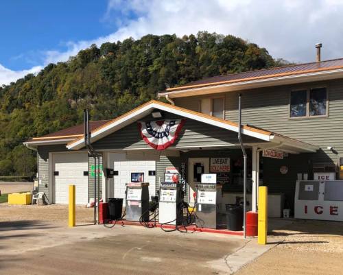 A small gas station with a green building, gas pumps, and patriotic decorations, set against a wooded hillside.