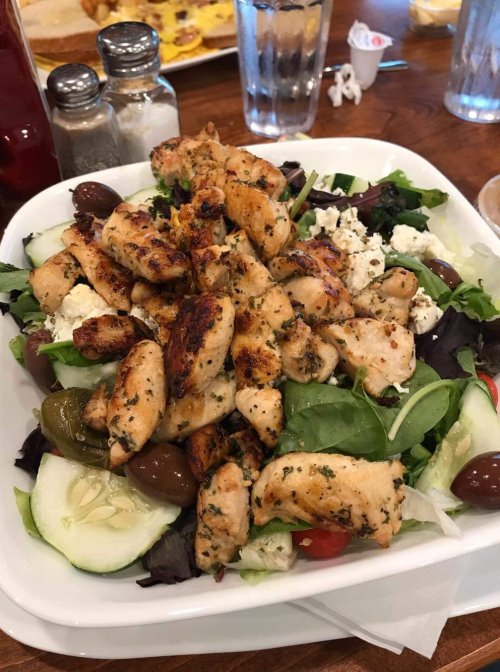 A fresh salad topped with grilled chicken, olives, cucumbers, and feta cheese on a white plate.