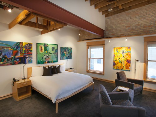 A modern bedroom with a large bed, colorful artwork on the walls, and cozy seating areas under wooden beams.
